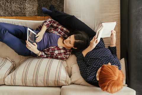 Erwachsene Tochter benutzt Tablet und Mutter liest zu Hause ein Buch, lizenzfreies Stockfoto