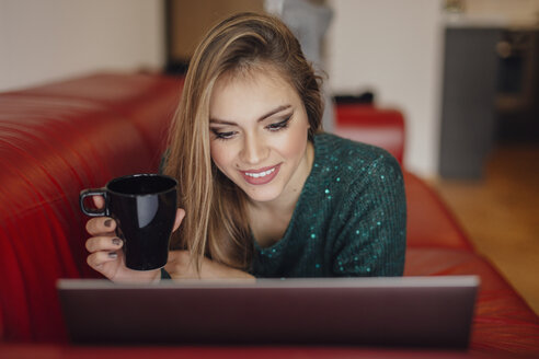 Junge Frau entspannt sich auf der Couch mit einer Tasse Kaffee und einem Laptop - LCUF00107