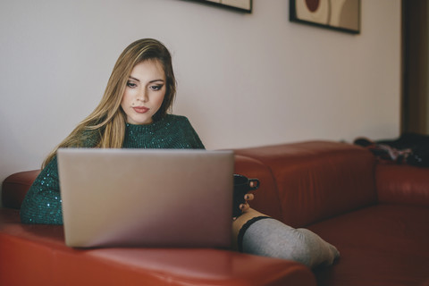 Junge Frau sitzt mit einer Tasse Kaffee auf der Couch und benutzt einen Laptop, lizenzfreies Stockfoto
