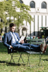 Businessman in Manhattan sitting on garden chair using digital tablet with feet up - GIOF02063