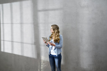Blonde Frau mit Tablet vor einer grauen Wand, die in die Ferne schaut - FMKF03562