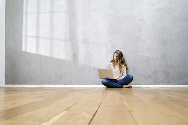 Blonde Frau sitzt auf dem Boden vor einer grauen Wand und benutzt einen Laptop - FMKF03564