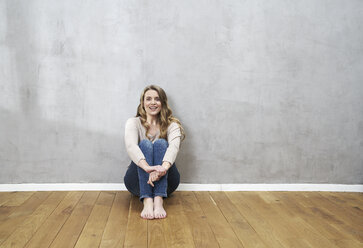 Lächelnde blonde Frau sitzt auf dem Boden vor einer grauen Wand - FMKF03565