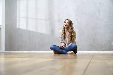 Laughing blond woman sitting on the floor with cell phone - FMKF03567