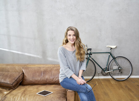 Porträt einer lächelnden blonden Frau, die zu Hause auf einer Ledercouch sitzt - FMKF03570