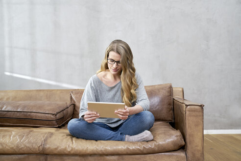 Blonde Frau sitzt auf einer Ledercouch und benutzt ein Tablet - FMKF03572