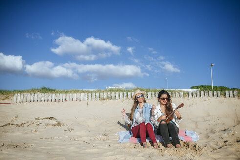 Zwei junge Frauen mit Ukulele sitzen auf einer Decke am Strand - KIJF01306