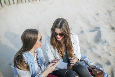 Zwei junge Frauen sitzen am Strand und benutzen ihr Handy - KIJF01310