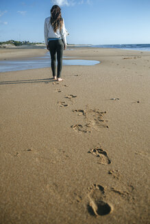 Back view of young woman walking on the beach leaving her footprints in the sand - KIJF01311