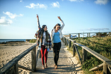 Two happy young woman walking barefoot on boardwalk - KIJF01324