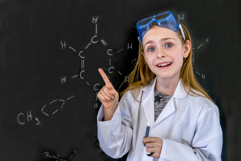 Porträt eines Mädchens mit Arbeitskittel und Schutzbrille vor einer Tafel mit chemischen Formeln, lizenzfreies Stockfoto