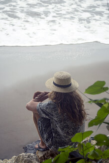 Indonesia, Java, woman wearing straw hat sitting on rock on the beach looking to the sea - KNTF00660