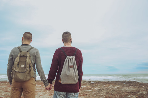 Rückenansicht eines jungen schwulen Paares mit Rucksäcken, das Hand in Hand am Strand steht und auf das Meer schaut, lizenzfreies Stockfoto