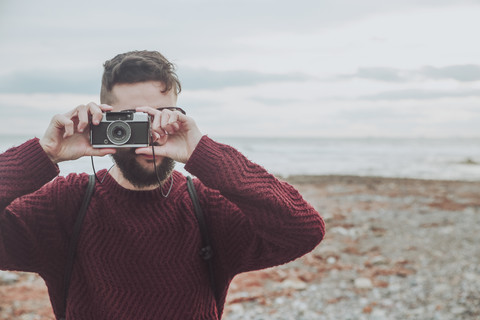Bärtiger Mann fotografiert am Strand mit einer alten Kamera, lizenzfreies Stockfoto