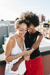 Female friends standing on rooftop, embracing and holding smart phones - GIOF02113