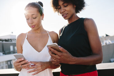 Female friends standing on rooftop using their smart phones - GIOF02115