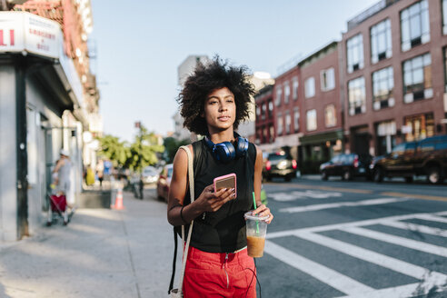 Junge Frau mit Kopfhörern und Smartphone beim Überqueren einer Straße in Brooklyn, mit einem Getränk zum Mitnehmen - GIOF02128