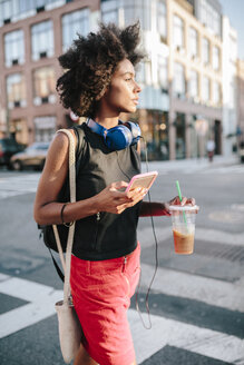 Junge Frau mit Kopfhörern und Smartphone beim Überqueren einer Straße in Brooklyn, mit einem Getränk zum Mitnehmen - GIOF02129