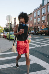 Junge Frau mit Kopfhörern und Smartphone beim Überqueren einer Straße in Brooklyn, mit einem Getränk zum Mitnehmen - GIOF02130