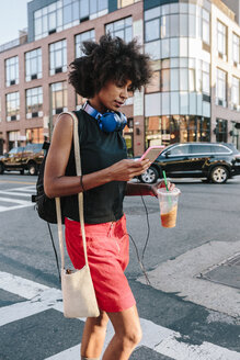 Junge Frau mit Kopfhörern und Smartphone beim Überqueren einer Straße in Brooklyn, mit einem Getränk zum Mitnehmen - GIOF02131