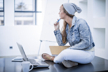 Young woman working in office sitting cross-legged on desk with her laptop - ZEF13117