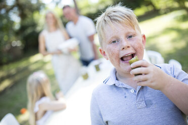 Portrait of boy eating gherkin outdoors with family in background - WESTF22817