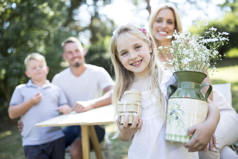 Porträt eines lächelnden Mädchens im Freien, das einen Krug und Kisten mit seiner Familie im Hintergrund hält - WESTF22819