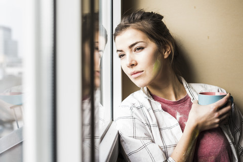 Junge Frau hält Kaffeetasse und schaut aus dem Fenster, lizenzfreies Stockfoto