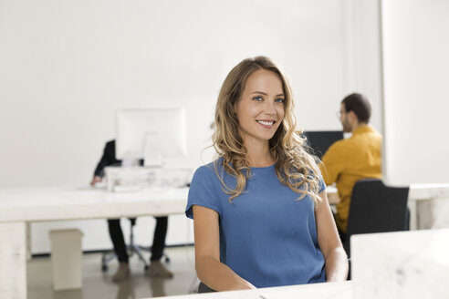 Lässige Geschäftsfrau sitzt im Büro, Kollegen arbeiten im Hintergrund - SBOF00354