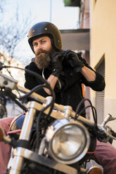 Porträt eines bärtigen Motorradfahrers, der auf seinem Motorrad sitzend seinen Helm befestigt - KKAF00491
