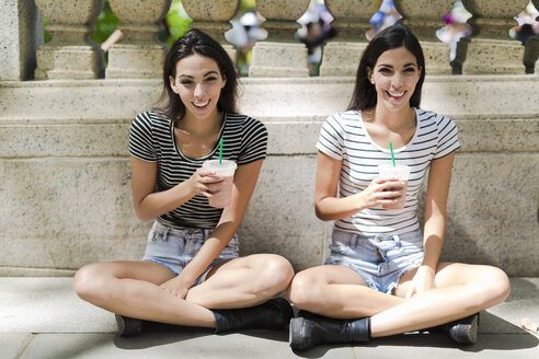 Zwei glückliche Zwillingsschwestern, die im Freien sitzen und ein Getränk zum Mitnehmen zu sich nehmen - GIOF02202