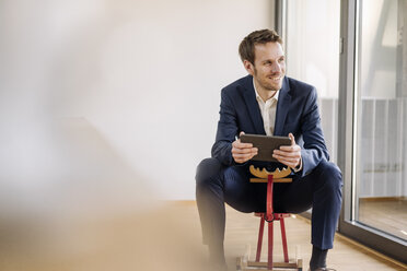 Smiling businessman sitting on rocking horse with tablet - KNSF01154