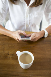 Frau mit Tasse Kaffee am Tisch, die ein Mobiltelefon benutzt - KNTF00780