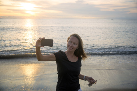 Indonesien, Bali, Frau macht ein Selfie am Strand bei Sonnenuntergang, lizenzfreies Stockfoto