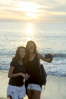 Indonesien, Bali, zwei Frauen machen ein Selfie am Strand bei Sonnenuntergang - KNTF00797
