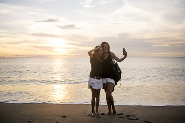 Indonesien, Bali, zwei Frauen machen ein Selfie am Strand bei Sonnenuntergang - KNTF00798