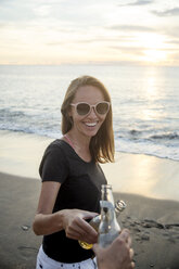 Indonesien, Bali, glückliche Frau stößt mit einem Freund am Strand bei Sonnenuntergang auf eine Bierflasche an - KNTF00800