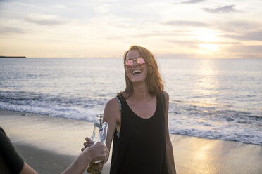 Indonesien, Bali, glückliche Frau stößt mit einem Freund am Strand bei Sonnenuntergang auf eine Bierflasche an - KNTF00801
