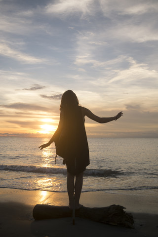 Indonesien, Bali, Frau beobachtet den Sonnenuntergang über dem Meer und balanciert auf einem Baumstamm, lizenzfreies Stockfoto