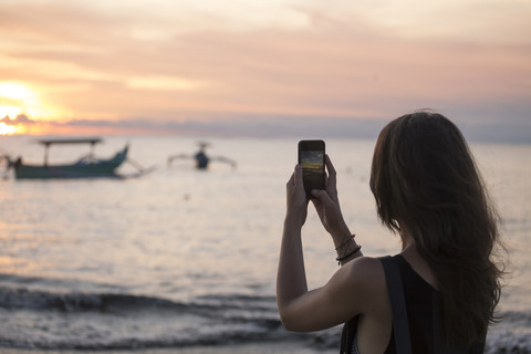 Indonesien, Bali, Frau beim Fotografieren des Sonnenuntergangs über dem Meer, lizenzfreies Stockfoto