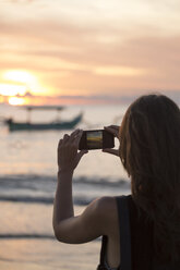 Indonesia, Bali, woman taking a picture of the sunset over the ocean - KNTF00805