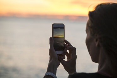 Indonesia, Bali, woman taking a picture of the sunset over the ocean - KNTF00810