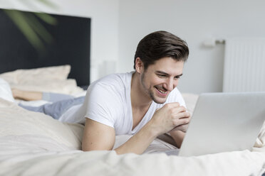 Lächelnder Mann auf dem Bett liegend mit Laptop - SHKF00756