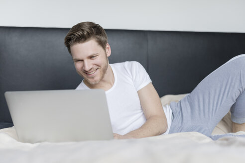 Lächelnder junger Mann auf dem Bett liegend mit Laptop - SHKF00757