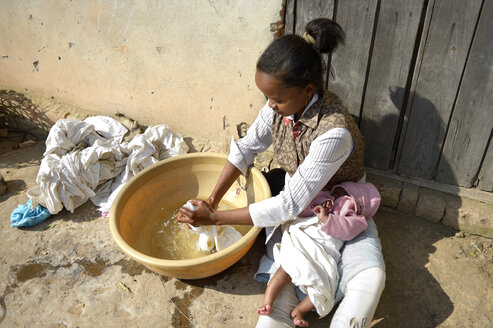 Madagaskar, Fianarantsoa, Junge Mutter mit Baby auf dem Schoß wäscht Wäsche in einer Schüssel - FLKF00797