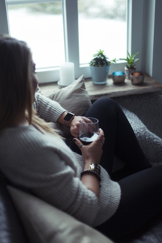 Junge Frau mit einem Glas Kaffee, die sich zu Hause auf der Couch entspannt und auf ihre Smartwatch schaut, lizenzfreies Stockfoto