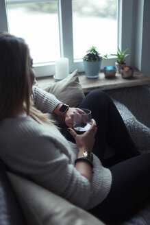 Young woman with glass of coffee relaxing on couch at home looking at smartwatch - NAF00059