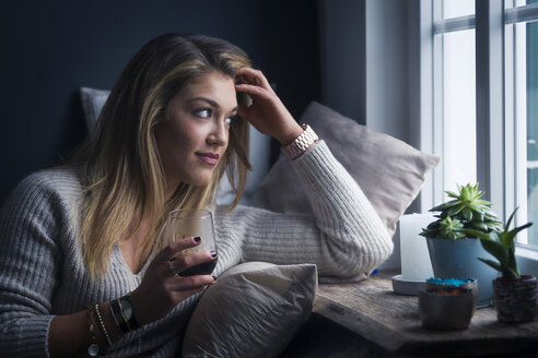 Young woman with glass of coffee sitting on couch at home looking through window - NAF00060