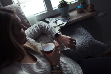 Junge Frau mit einem Glas Kaffee, die sich zu Hause auf der Couch entspannt und auf ihre Smartwatch schaut - NAF00063