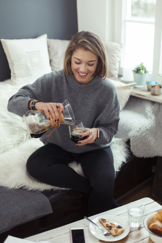 Lächelnde blonde Frau sitzt auf der Couch und gießt Kaffee in ein Glas, lizenzfreies Stockfoto